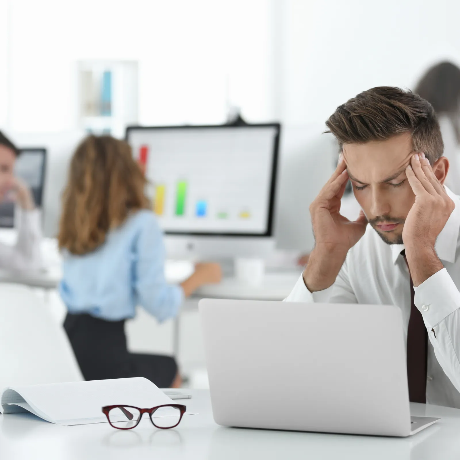 Stressed businessman holding his head while working on laptop in office with colleagues in background