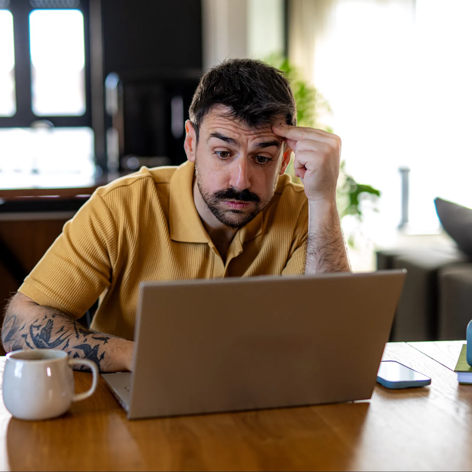 Man in yellow shirt focused and concerned working on laptop at wooden table with coffee and plant nearby.