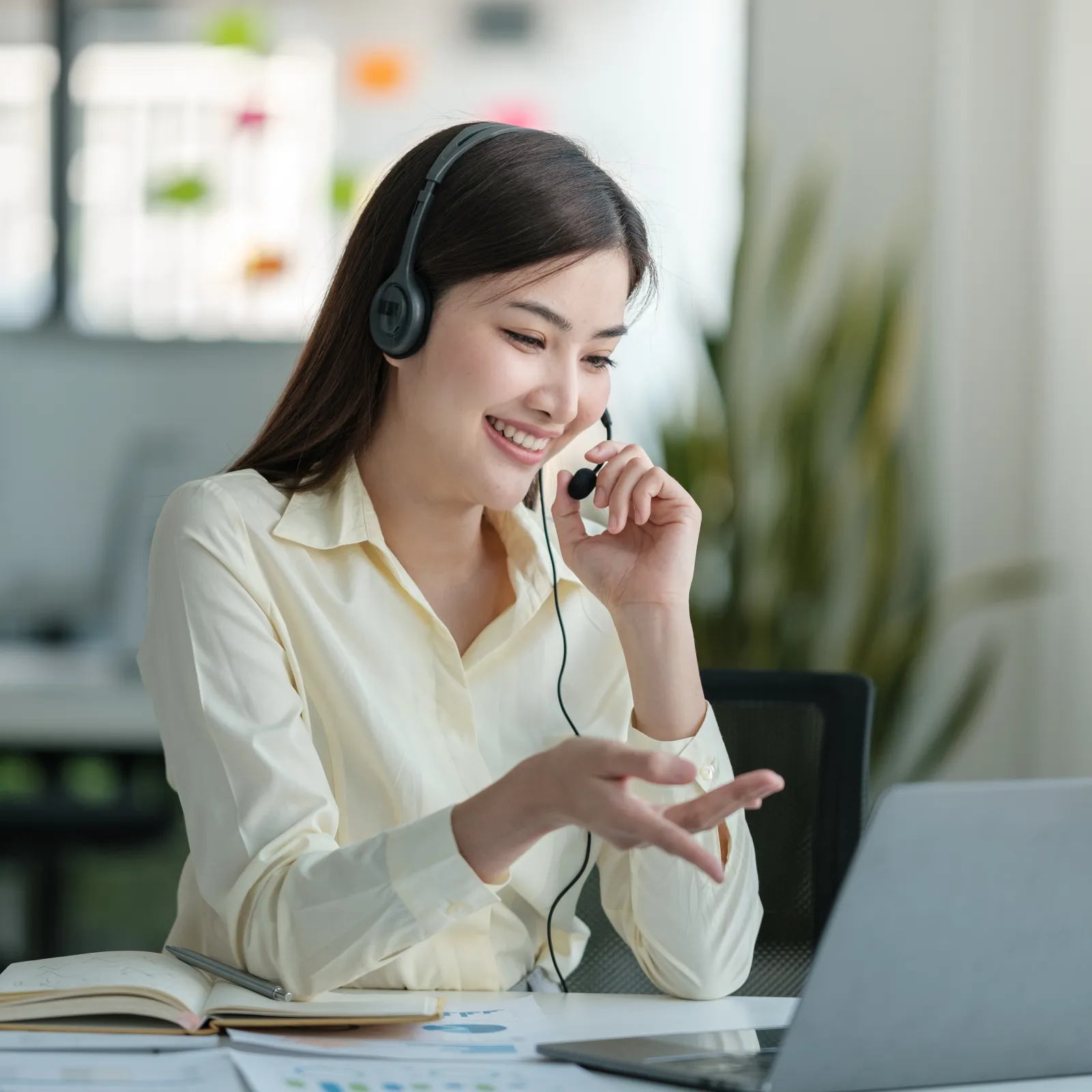 Smiling woman with headset working on laptop and taking notes in bright modern office environment