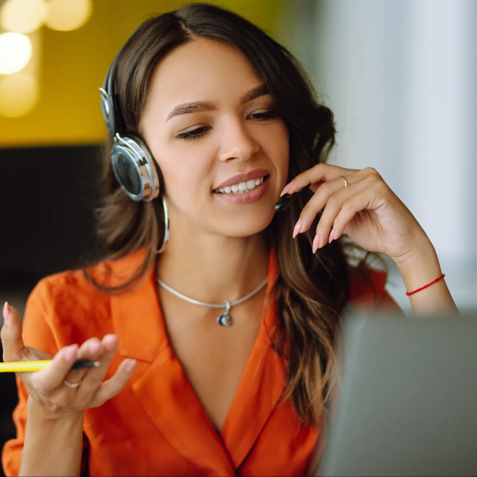 Woman in an orange blouse wearing headphones and speaking while looking at a laptop in a bright office.