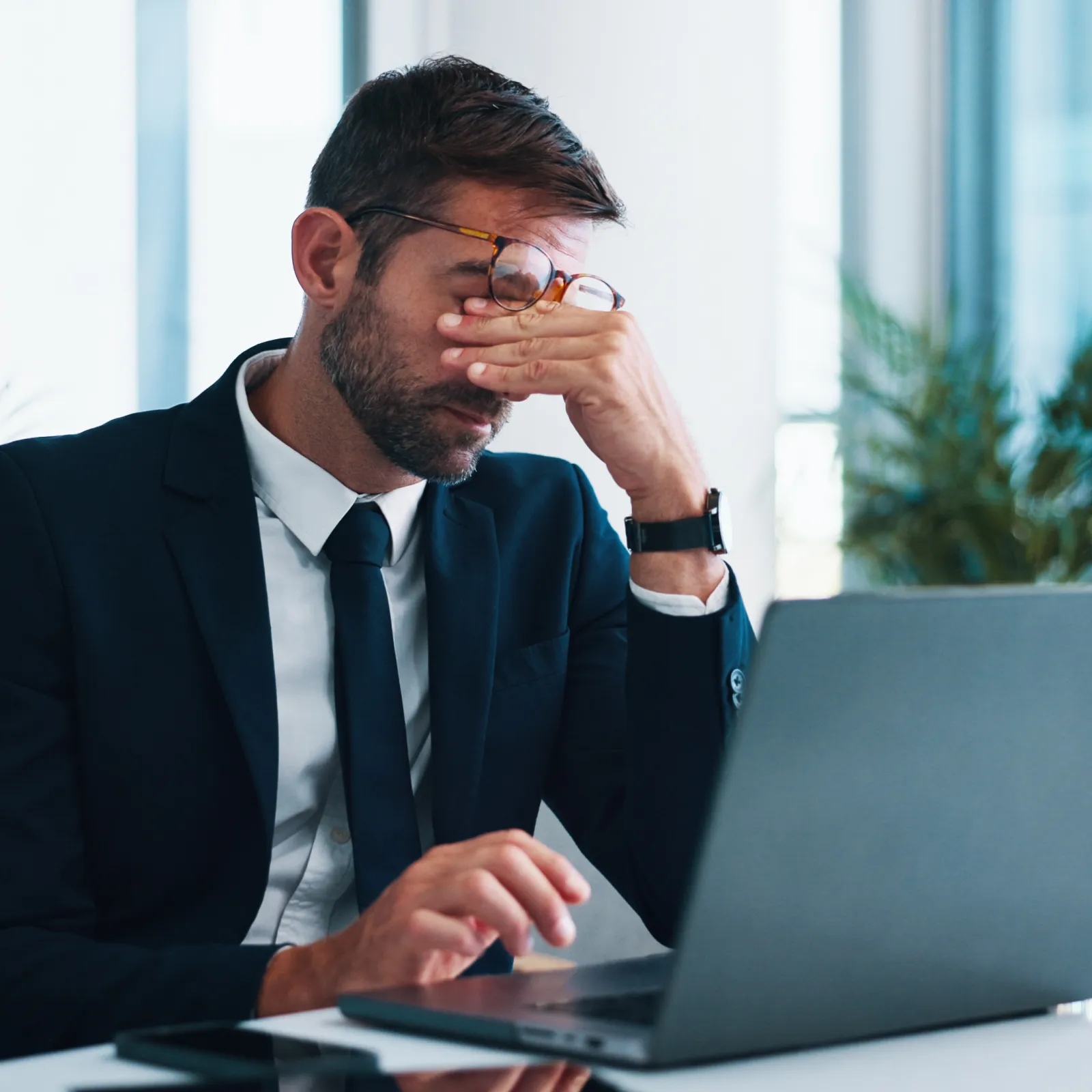 Stressed businessman in suit rubbing eyes while working on laptop in modern office with large windows.