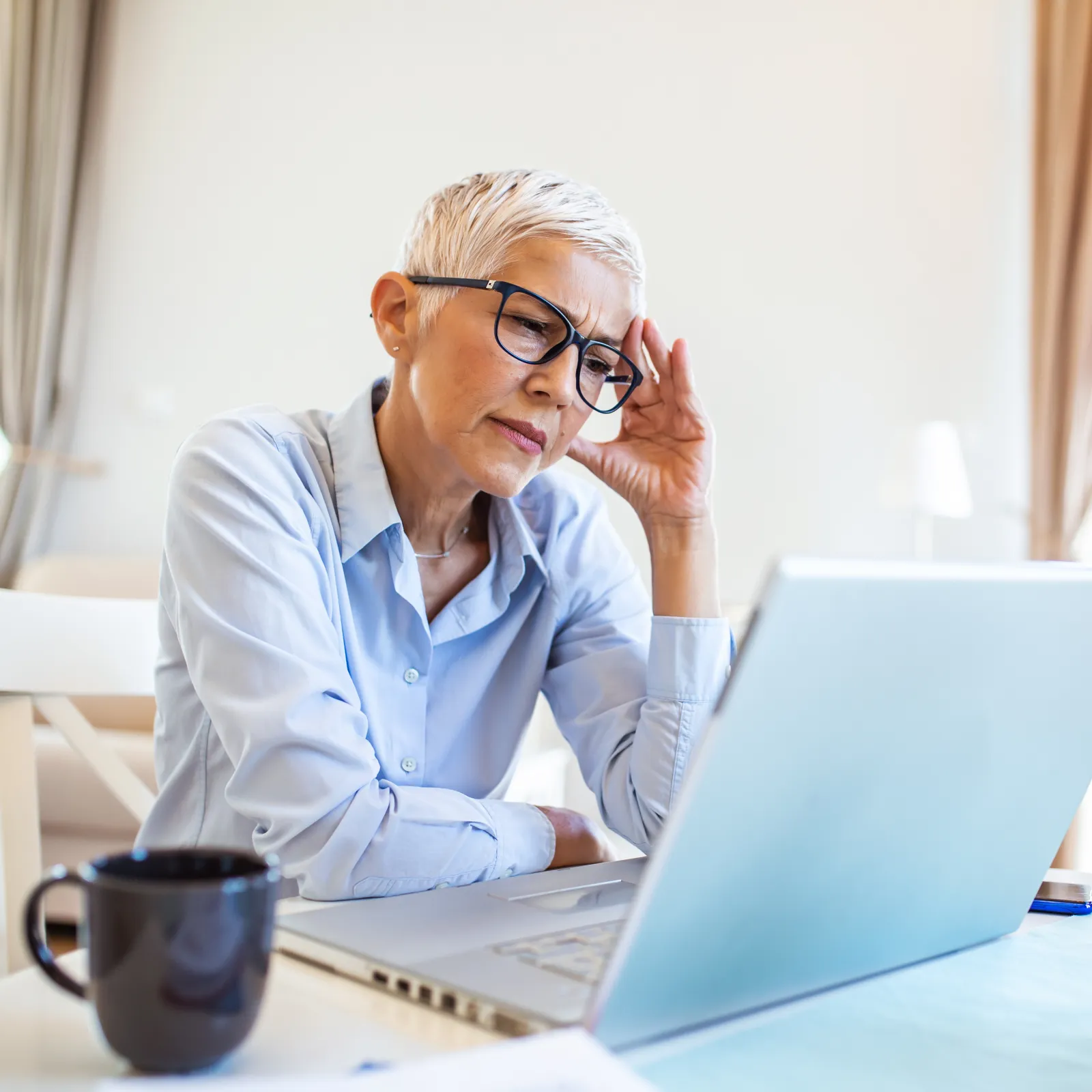 Senior woman in glasses working on laptop, looking concerned in bright modern home office with coffee and plant.