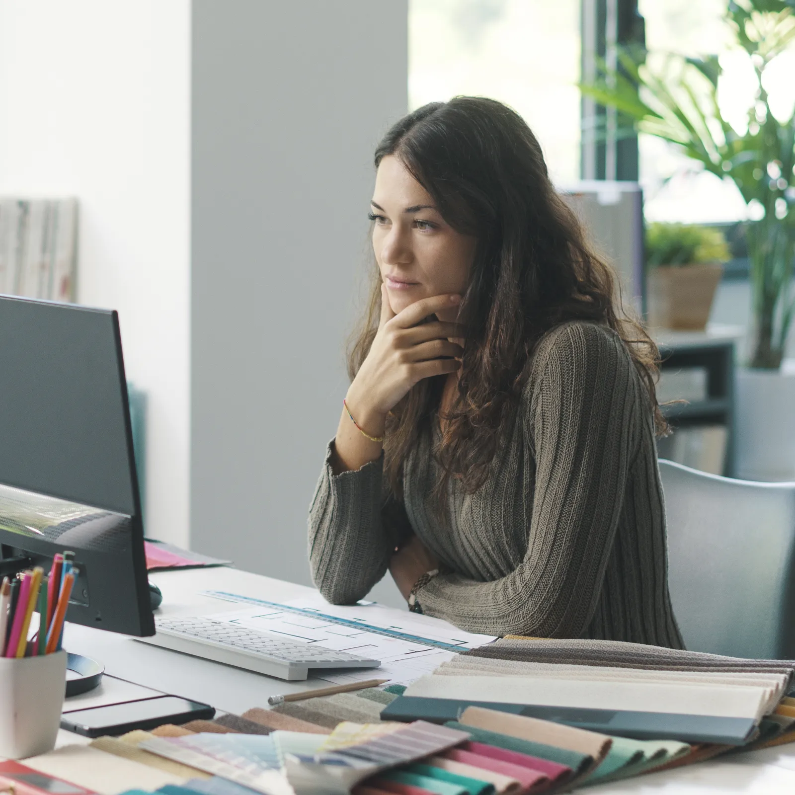 Woman thoughtfully working at a cluttered desk with computer, art supplies, and stacks of paper in a bright office.