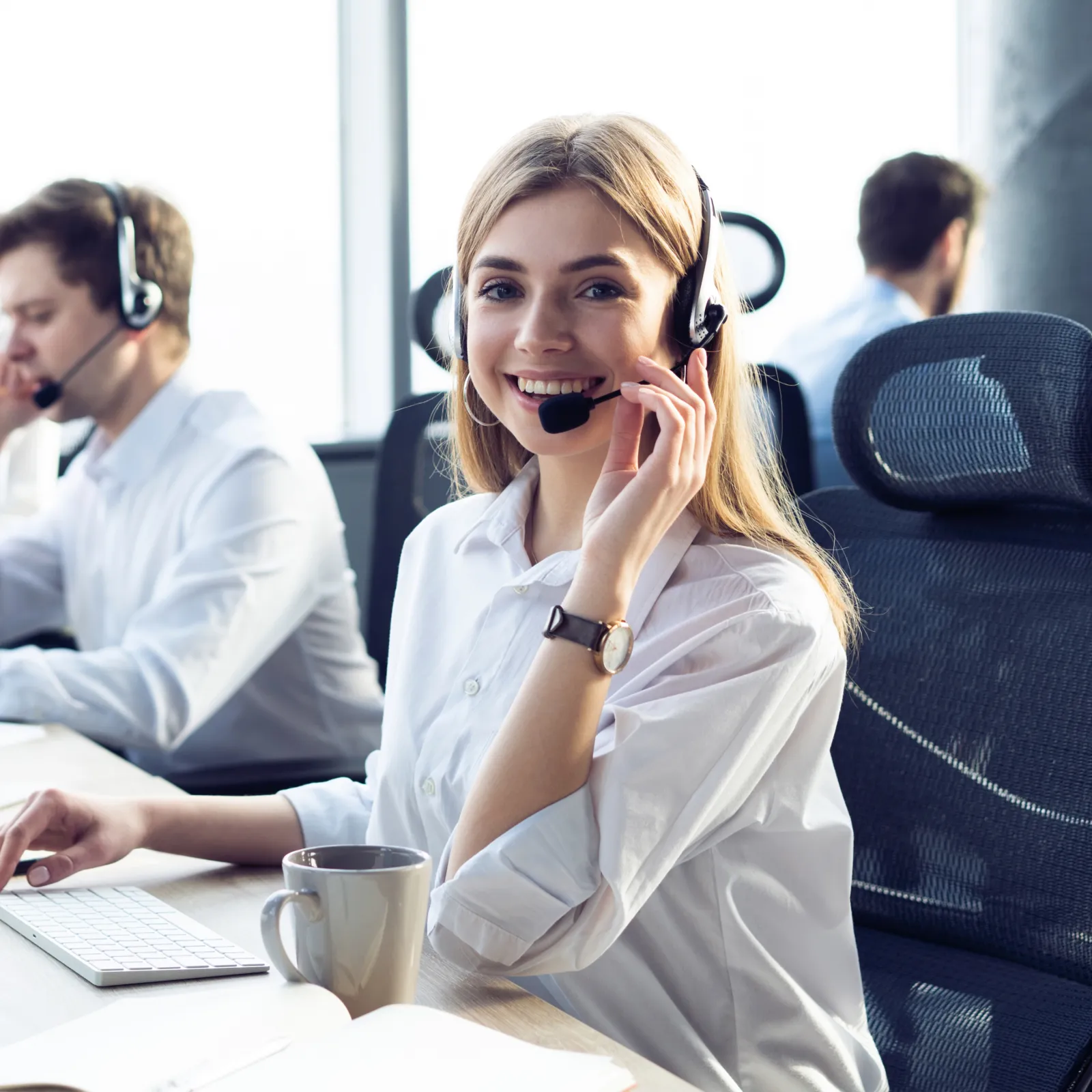 Smiling woman in headset working at computer in a bright call center office with colleagues in background.