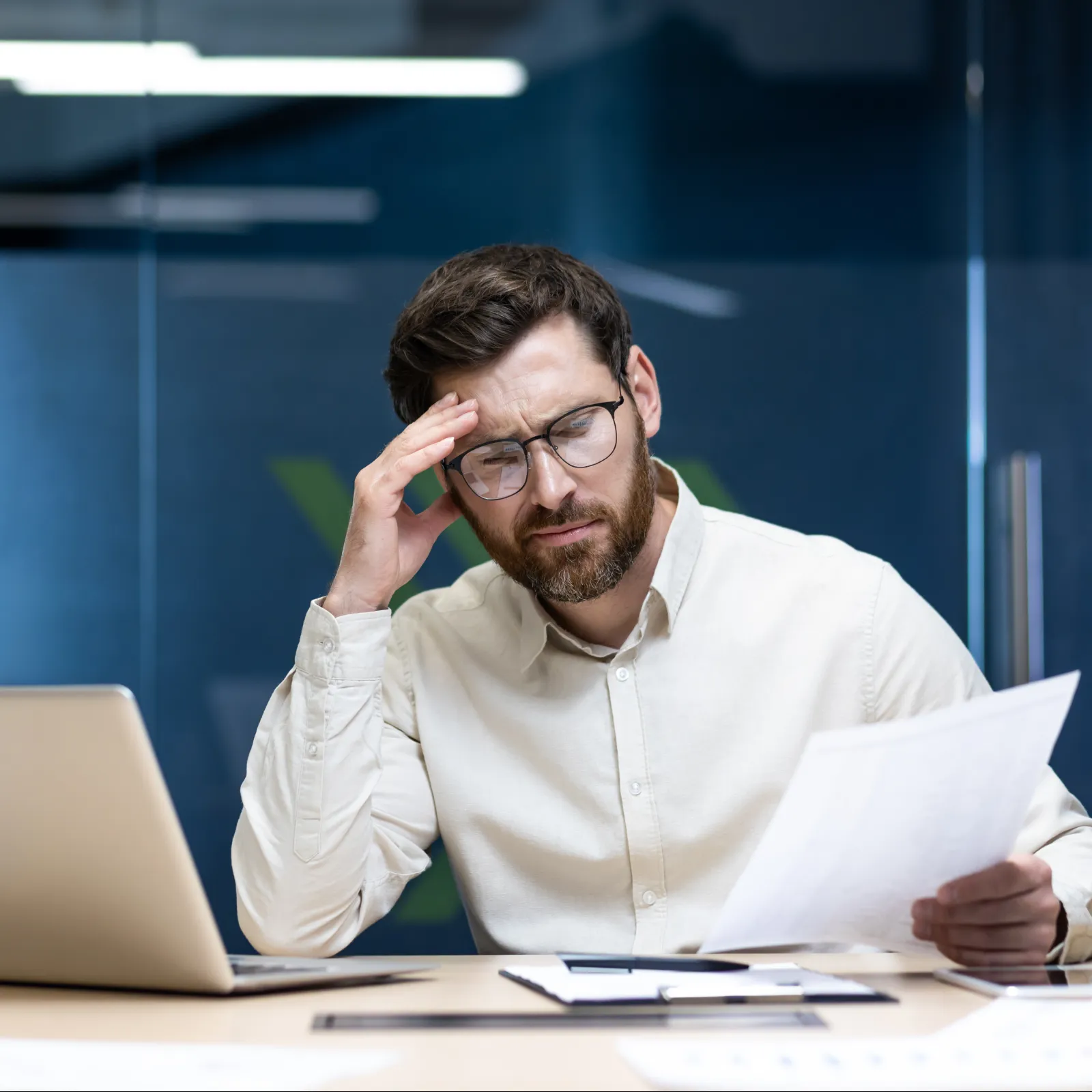 Stressed man in glasses holding papers and rubbing forehead while working on laptop at office desk.