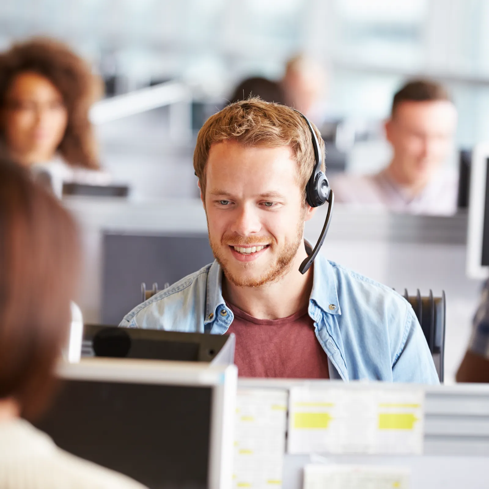 Smiling male customer service agent wearing headset working at his computer in a busy office environment.