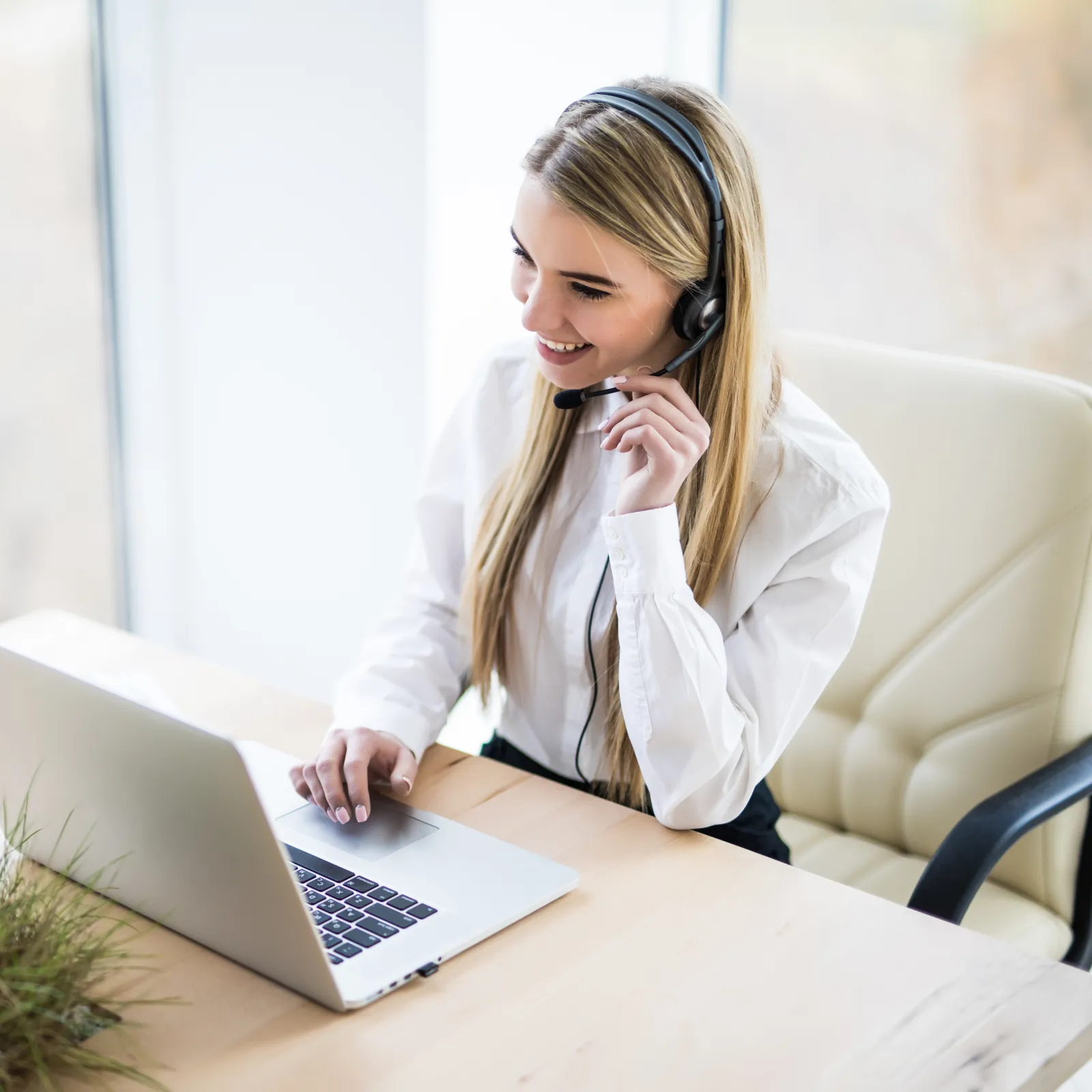 Young woman in headset smiling while working on laptop at modern office desk by window.
