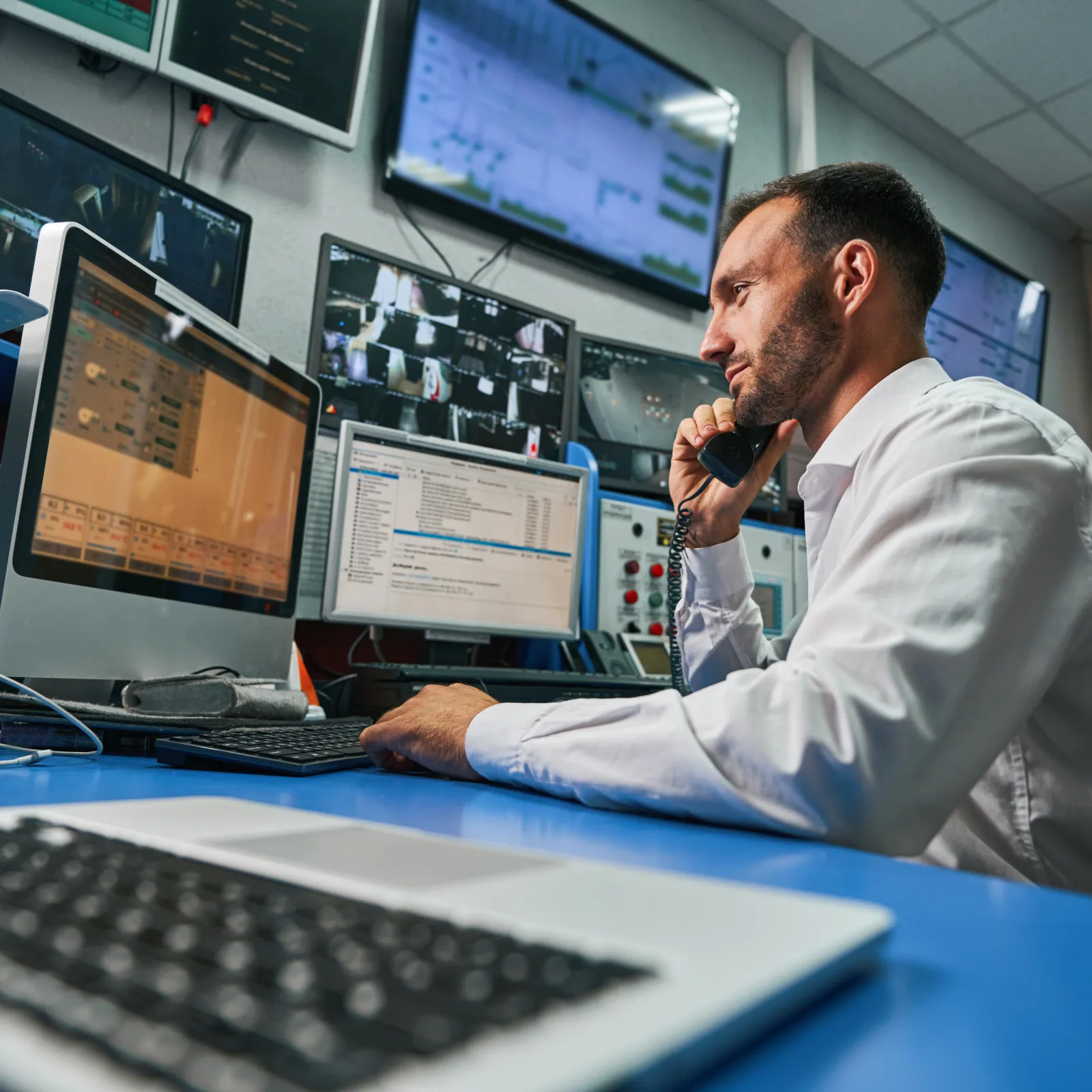 Man in control room monitoring security cameras and data on multiple screens while talking on a phone.
