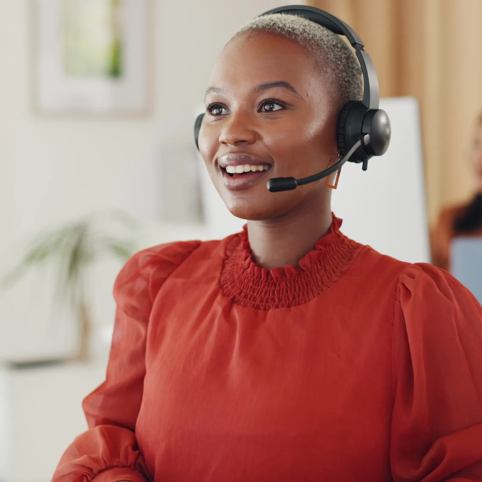 Smiling woman in red blouse wearing headset working at computer in modern office environment.
