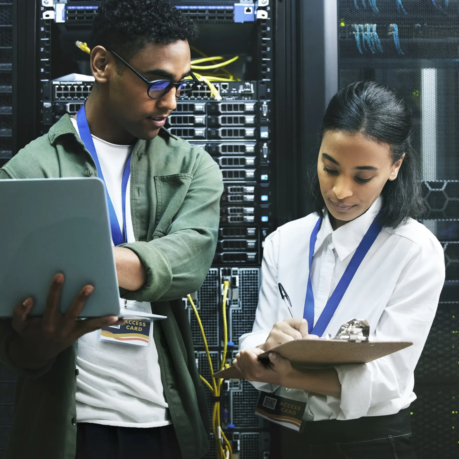 Two IT professionals working in a server room, one using a laptop and the other taking notes on a clipboard.