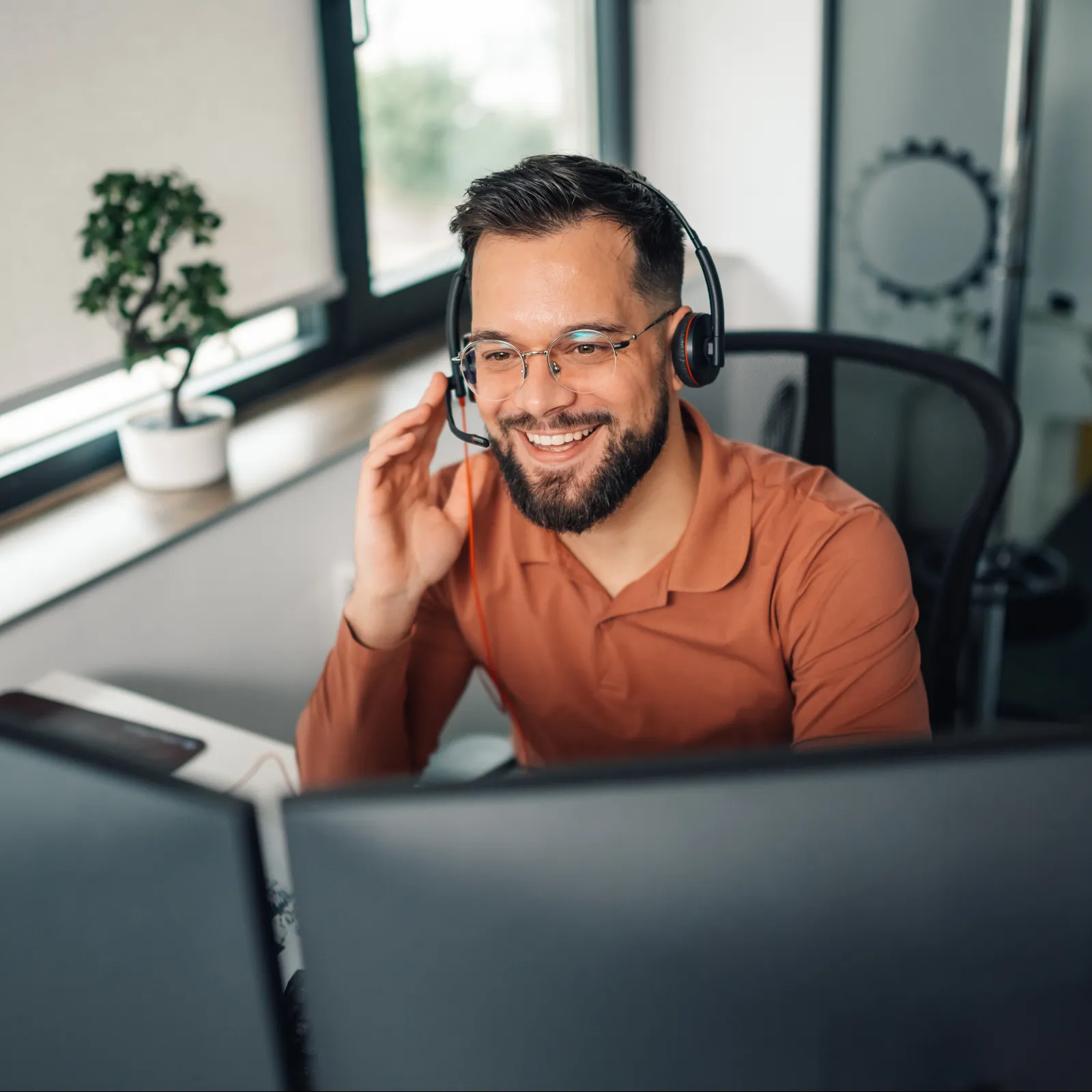 Smiling man with headset working at dual monitors in modern office with natural light and green plant.