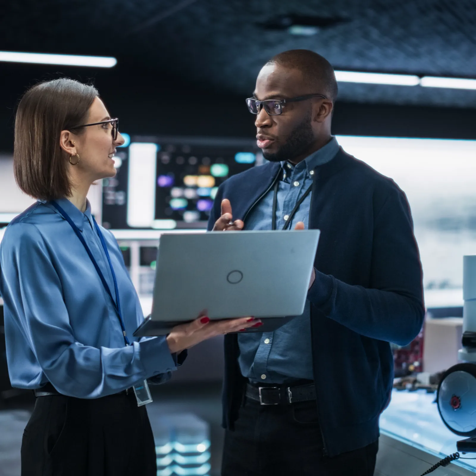 Two professionals discussing work while holding a laptop in a modern high-tech laboratory setting.