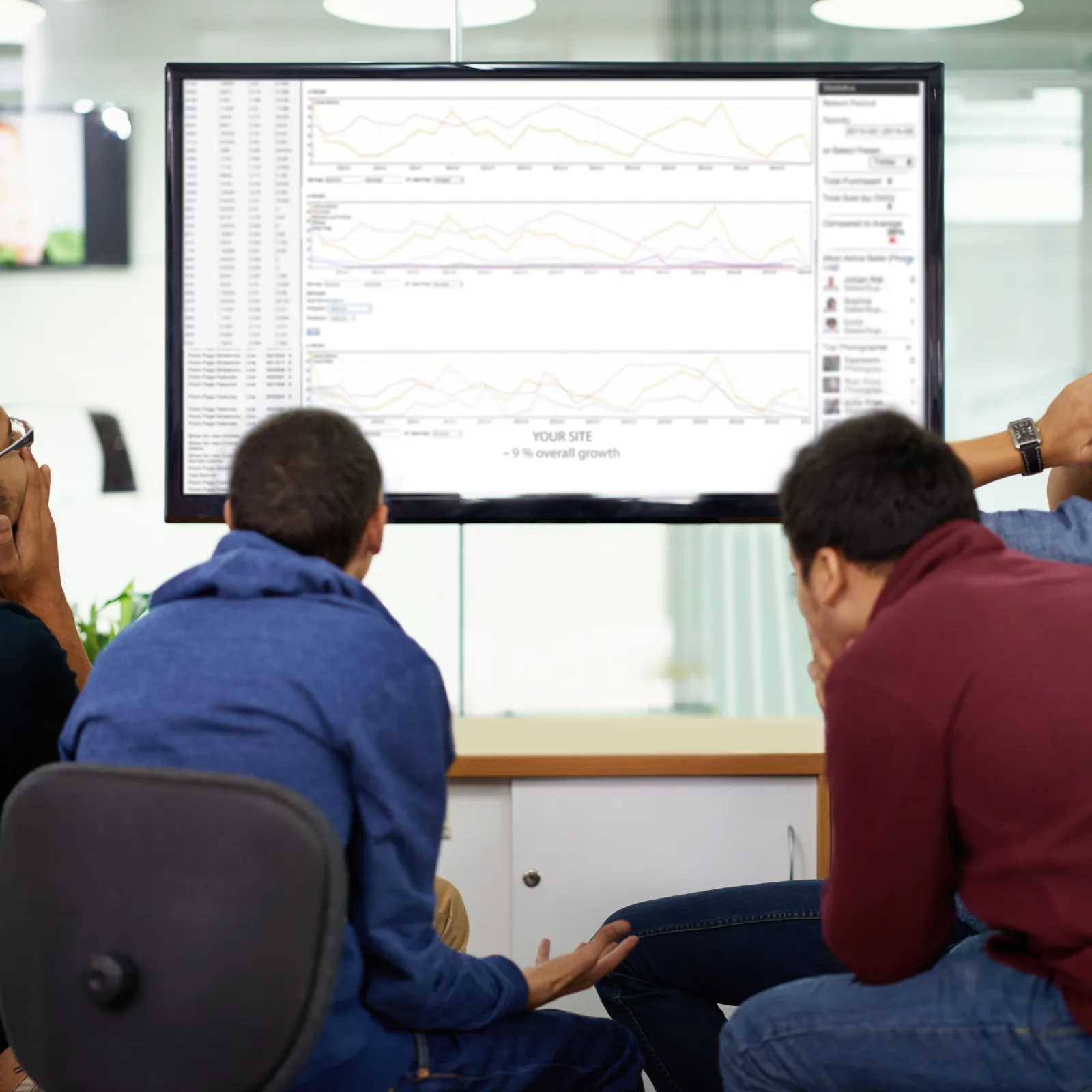 Four men sitting in office chairs looking at a large screen displaying charts and data analysis in a meeting room.