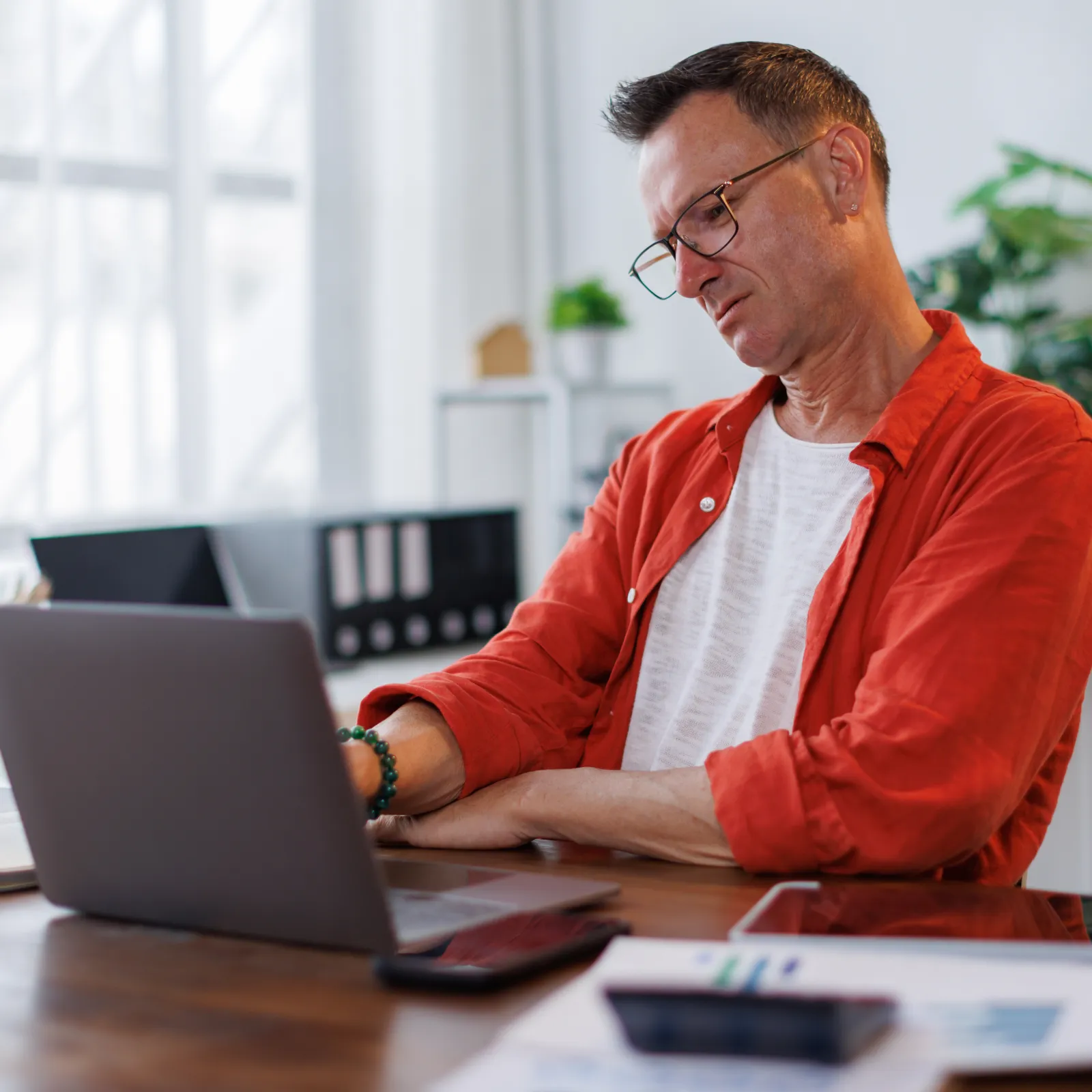 Man wearing glasses and an orange shirt working on a laptop at a wooden desk in a bright room.