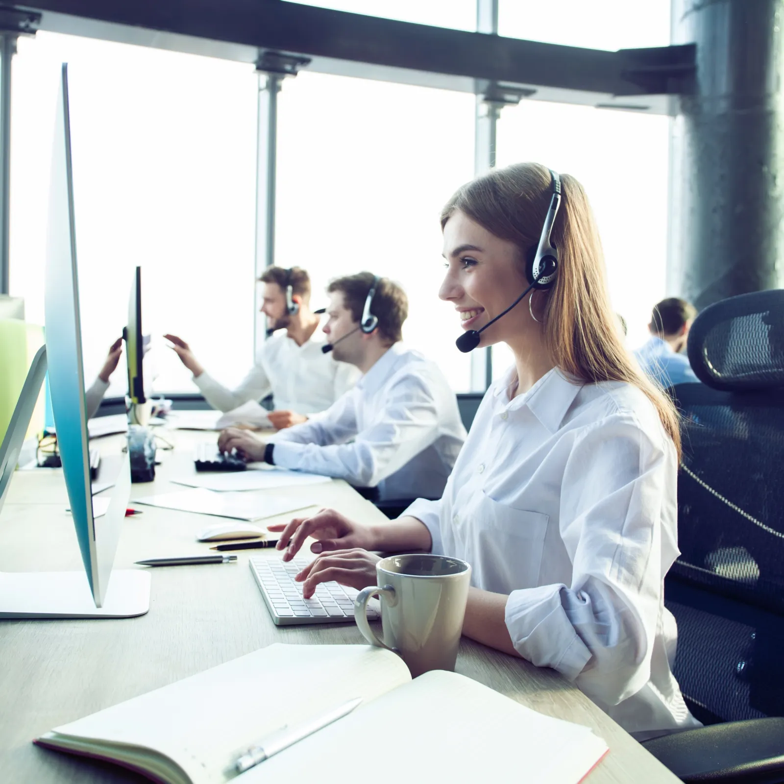 Smiling call center employees wearing headsets working on computers in a bright modern office space.