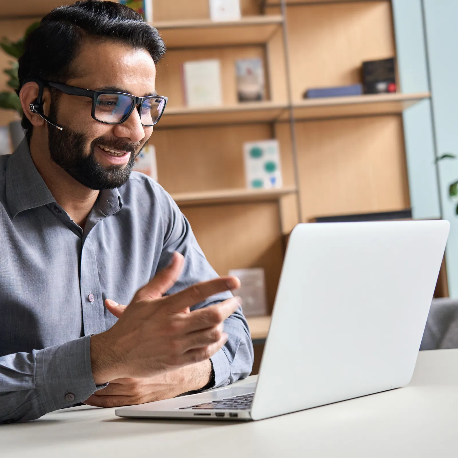 Smiling man wearing glasses and headset engaged in video call on laptop in modern office setting