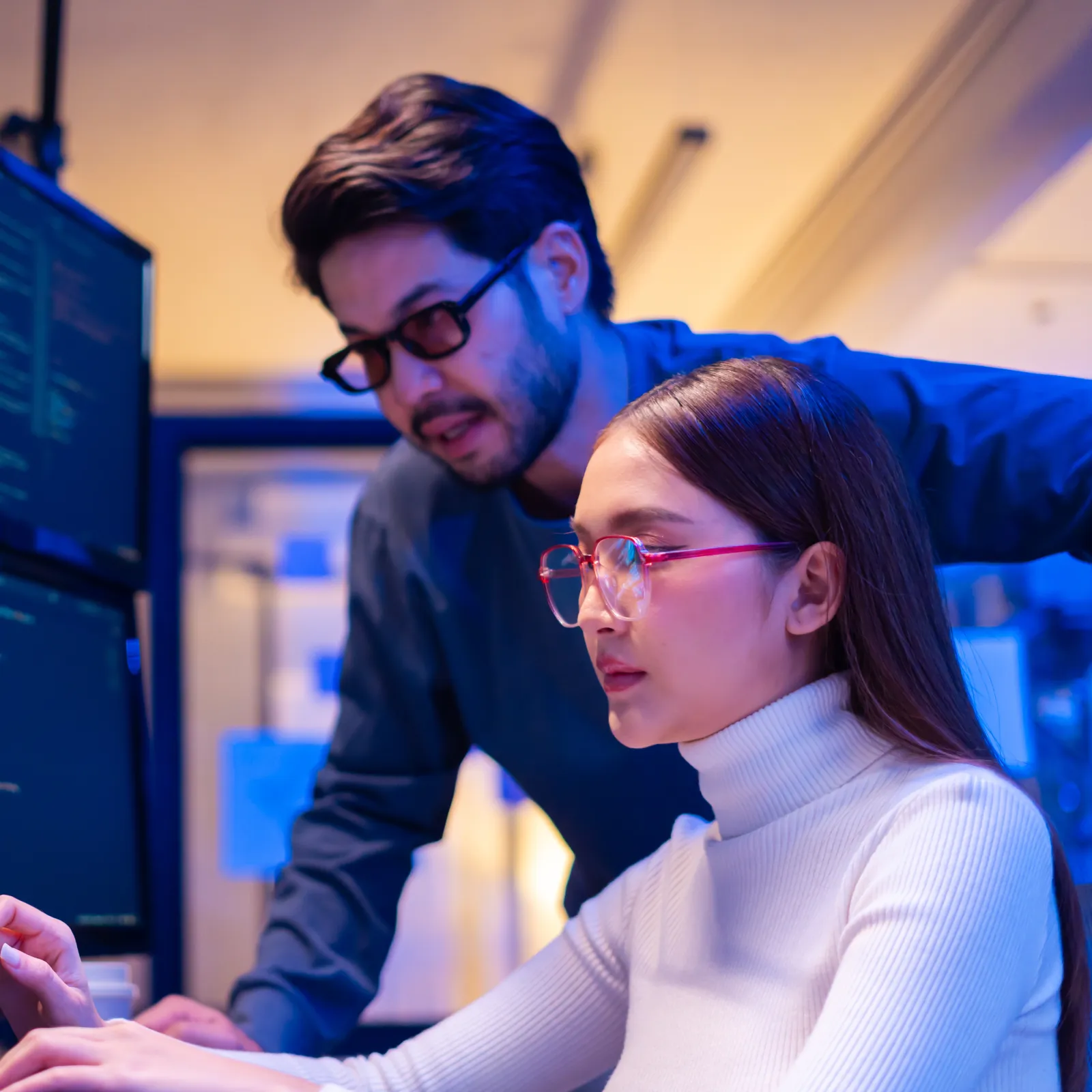 Two programmers collaborate over code on a multi-monitor setup in a modern office with blue ambient lighting.
