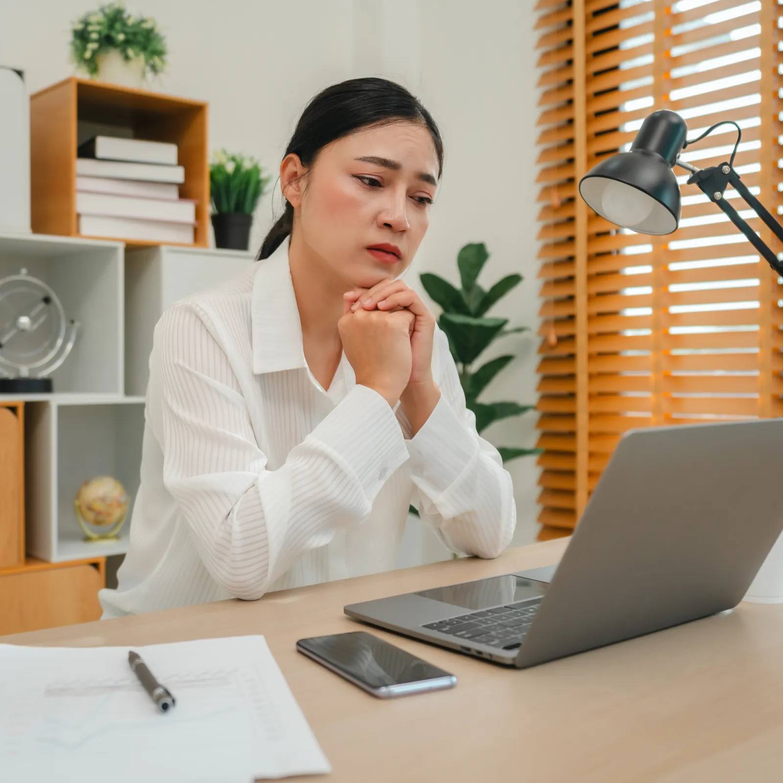 Worried woman in white shirt looking at laptop with smartphone and papers on desk in home office.