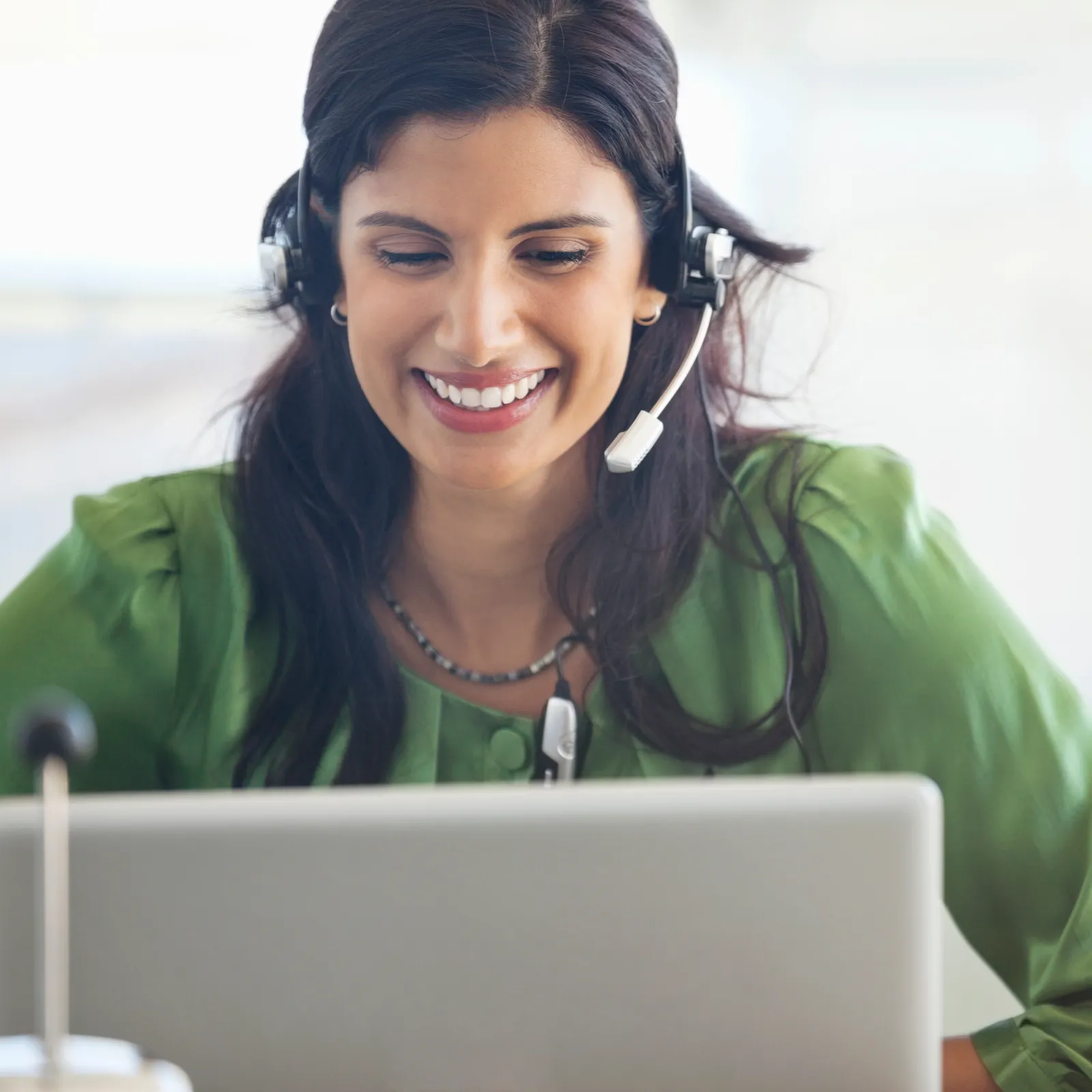 Smiling woman wearing headset and green blouse working on laptop in bright office environment.