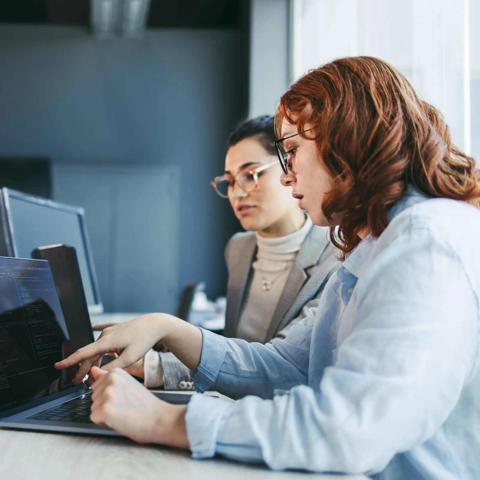 Two women collaborating on coding projects using a laptop in a modern office setting with natural light.