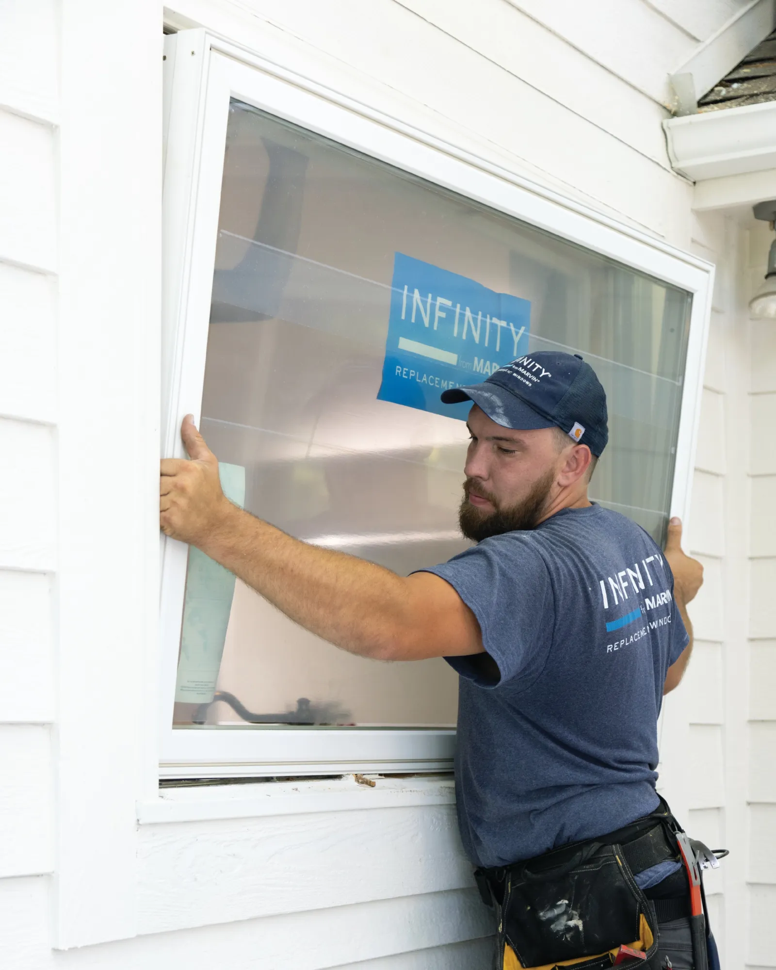 Worker wearing a blue shirt and cap installs a window in a white house exterior during daytime.