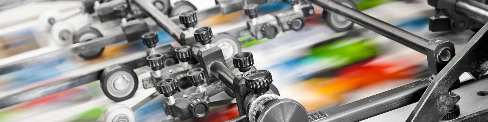 Close-up of a high-speed printing press producing colorful sheets with mechanical rollers and adjustment knobs.