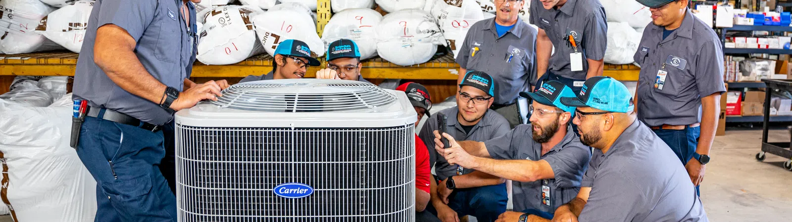 Group of technicians gathered around a Carrier HVAC unit inside a warehouse for training or inspection.