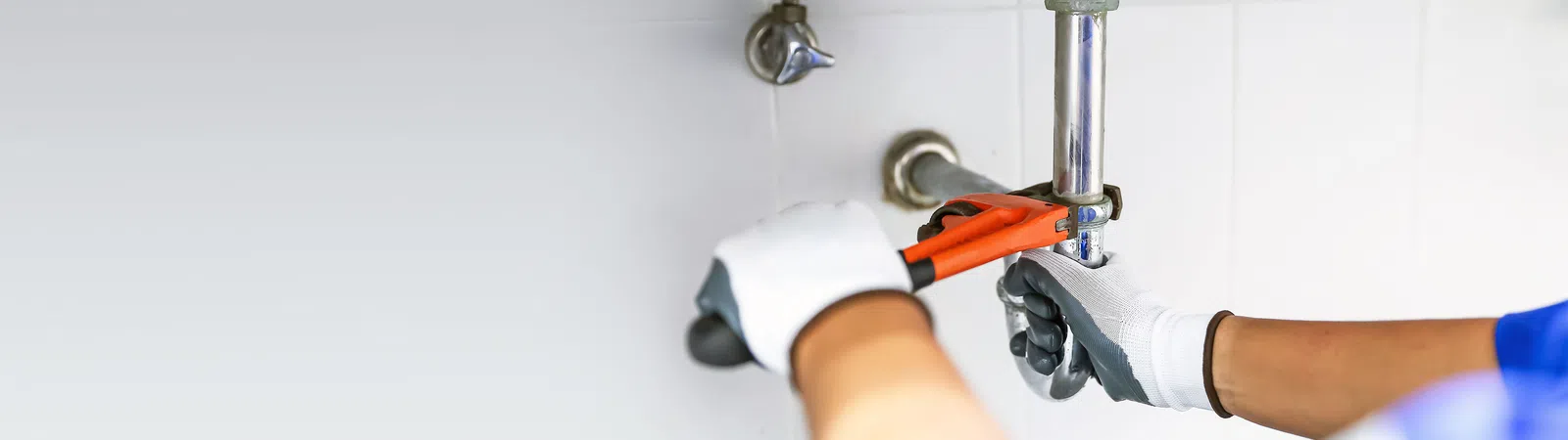 Plumber with gloves using a wrench to fix a pipe under a sink in a tiled bathroom or kitchen setting.