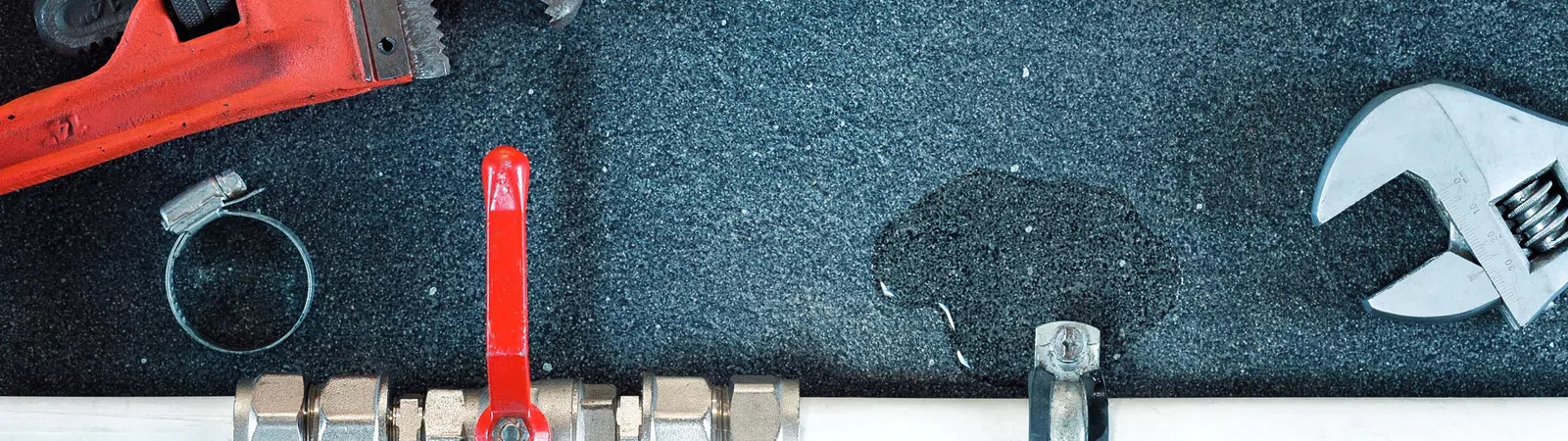 Plumbing tools and fittings including pipe wrench, adjustable wrench, valve, and PVC pipes on a wet surface.