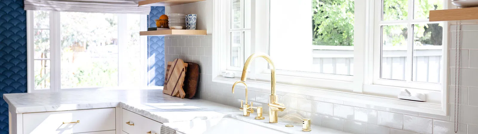 Bright modern kitchen with white marble countertops, gold fixtures, farmhouse sink, and large windows with natural light.