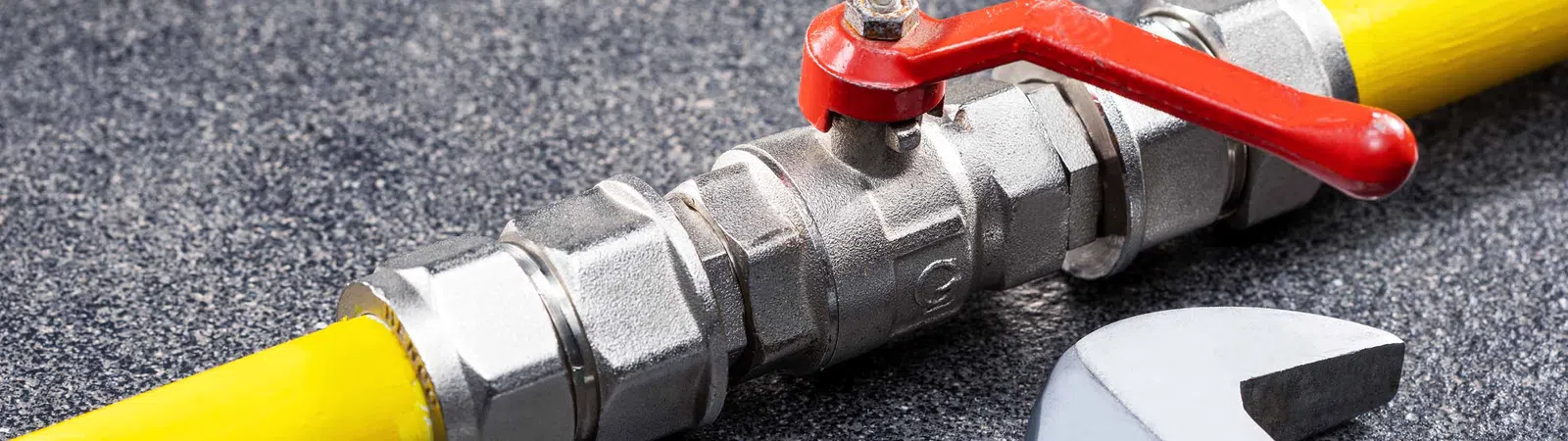 Close-up of a yellow gas pipe with a metal valve and red handle beside an adjustable wrench on a gray surface.