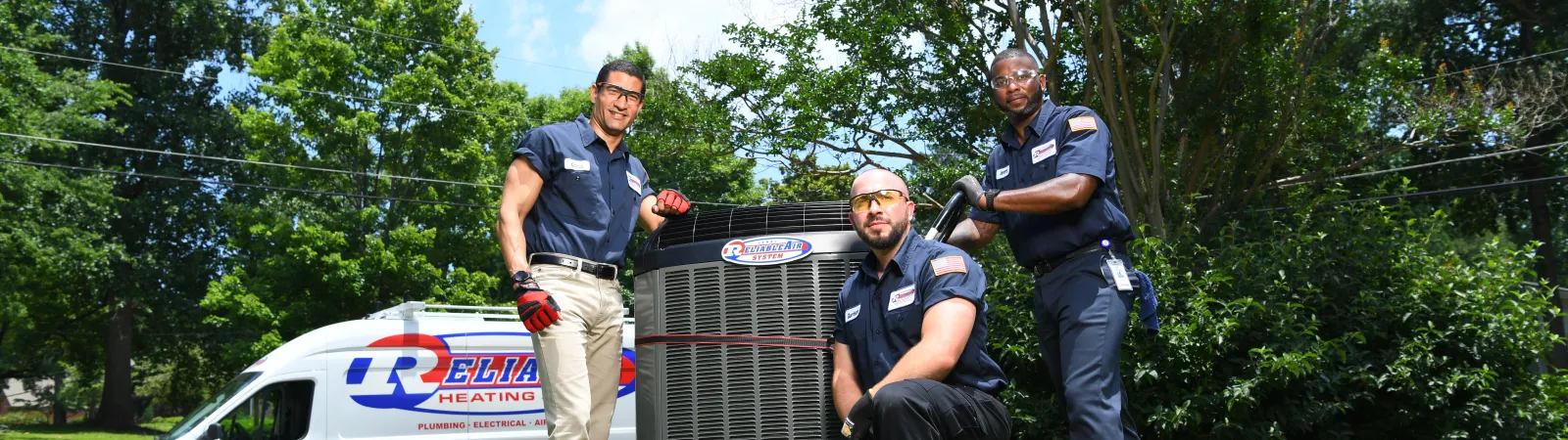 Three HVAC technicians posing with an air conditioning unit outdoors on green grass with a company van in the background