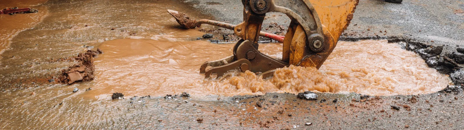 Excavator digging in a flooded street with muddy water rushing from a broken pipe.