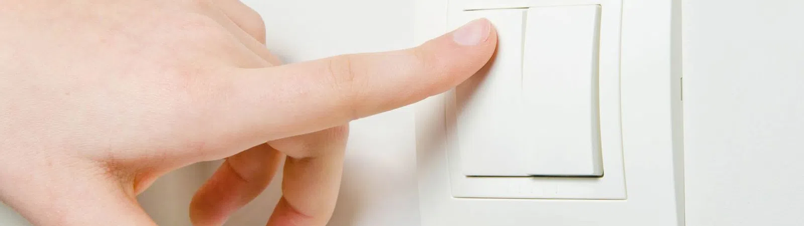 A person's finger pressing a light switch on a white wall, illuminating a room.
