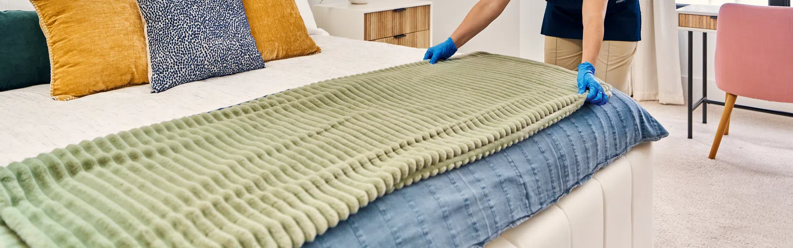 Housekeeper wearing gloves neatly making a modern bed with colorful pillows and green blanket in bright bedroom.