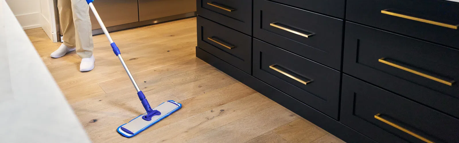 Woman wearing gloves mopping wood floor in modern kitchen with black cabinets and stainless steel fridge