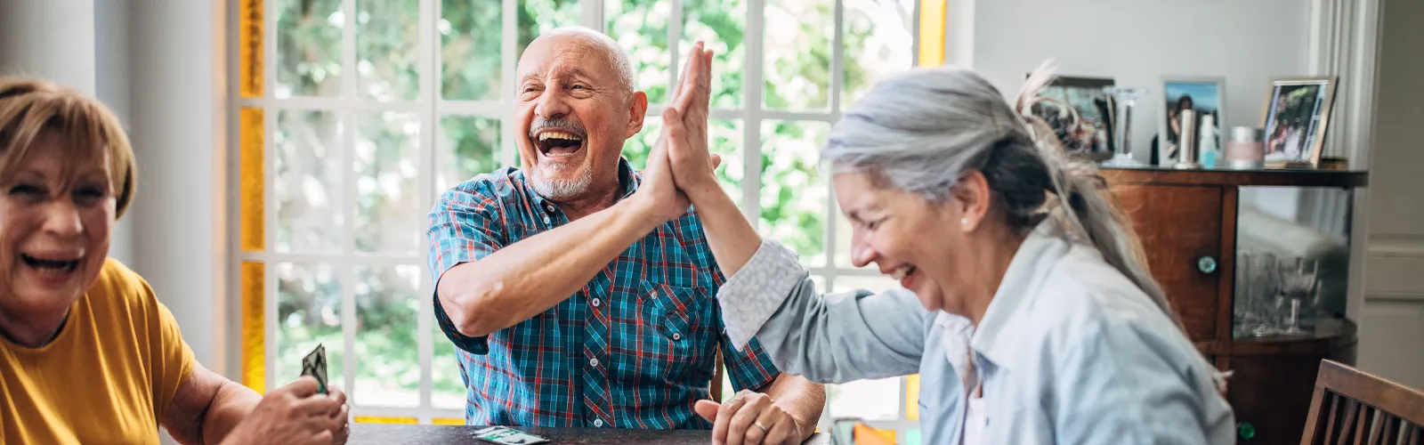 Happy elderly friends playing cards and sharing a joyful high five around a table in a bright room.