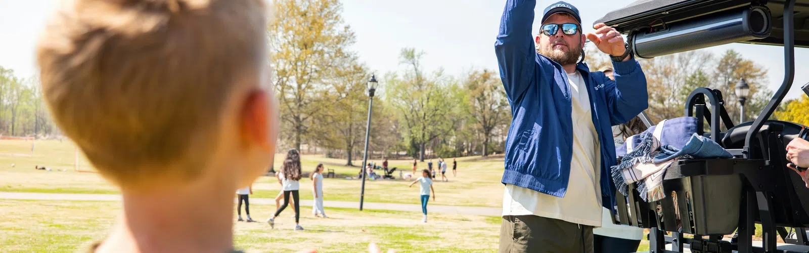 Man throwing volleyball to a child in a sunny park near a golf cart with people in the background