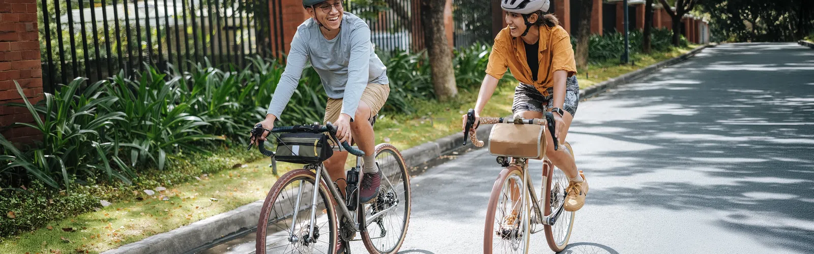 Two people wearing helmets smiling and riding bicycles on a sunny suburban street with greenery and houses.