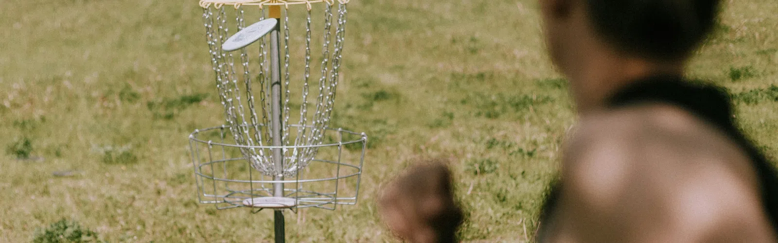Disc golf basket with a disc flying toward it on a grassy field, person blurred in foreground preparing to throw.