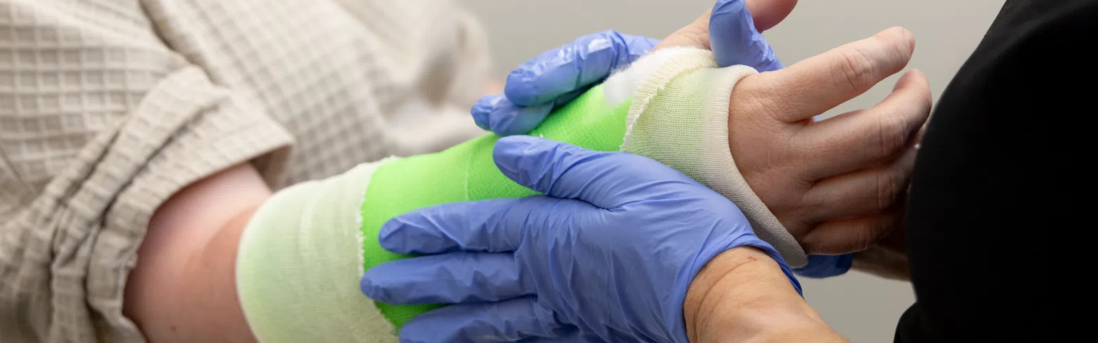 Healthcare professional wearing blue gloves adjusts green arm cast on patient in beige shirt.