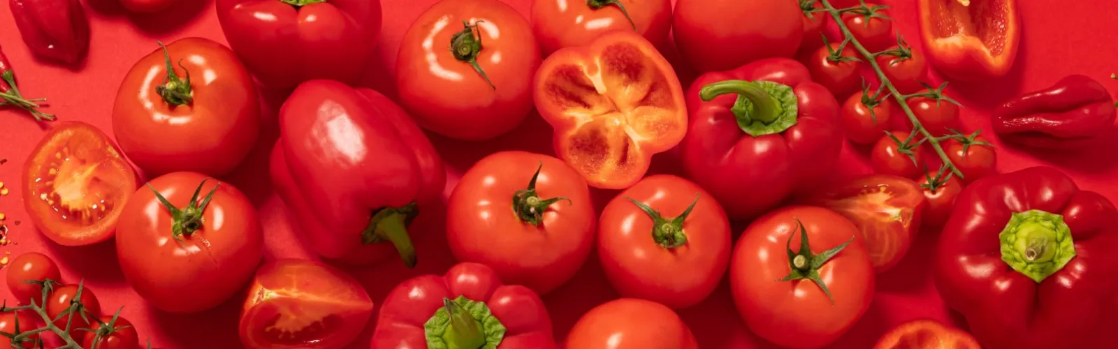 Assortment of red tomatoes, bell peppers, chili peppers, and crushed red pepper flakes on a red background.