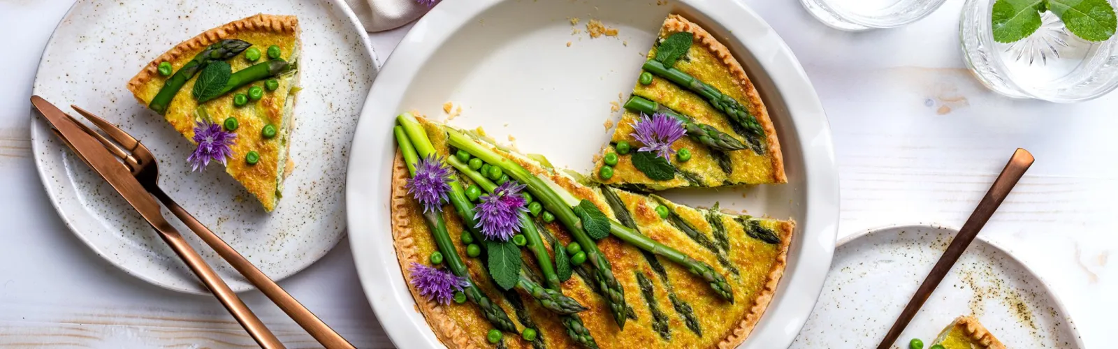 Asparagus and pea quiche garnished with purple flowers and fresh herbs served on white plates with copper utensils.
