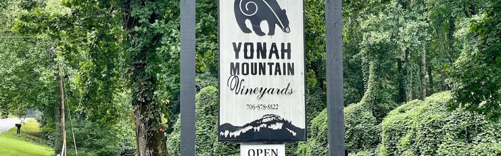 Wooden sign for Yonah Mountain Vineyards hangs between stone pillars with barrels at green forest roadside.