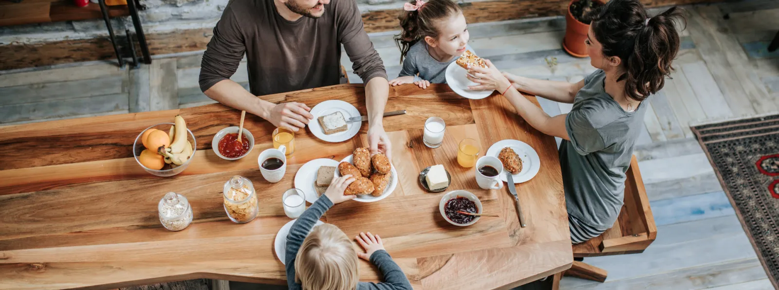 Family of four enjoying breakfast with pastries, juice, and coffee at wooden table in cozy home setting