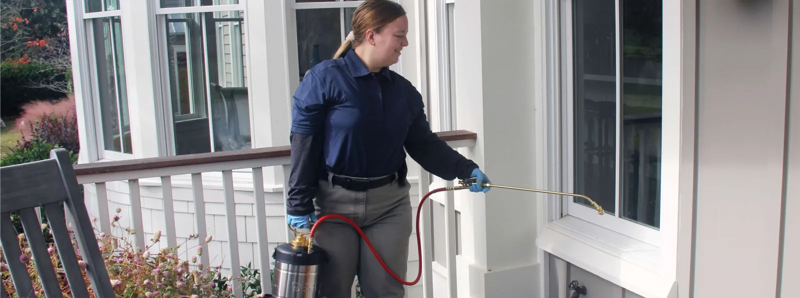 Woman in uniform spraying pesticide on house exterior near window for pest control service.