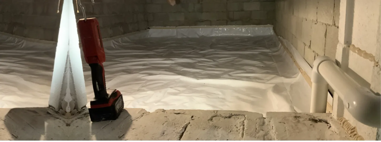 Basement with plastic vapor barrier installed on floor and a red cordless drill standing upright on concrete surface.