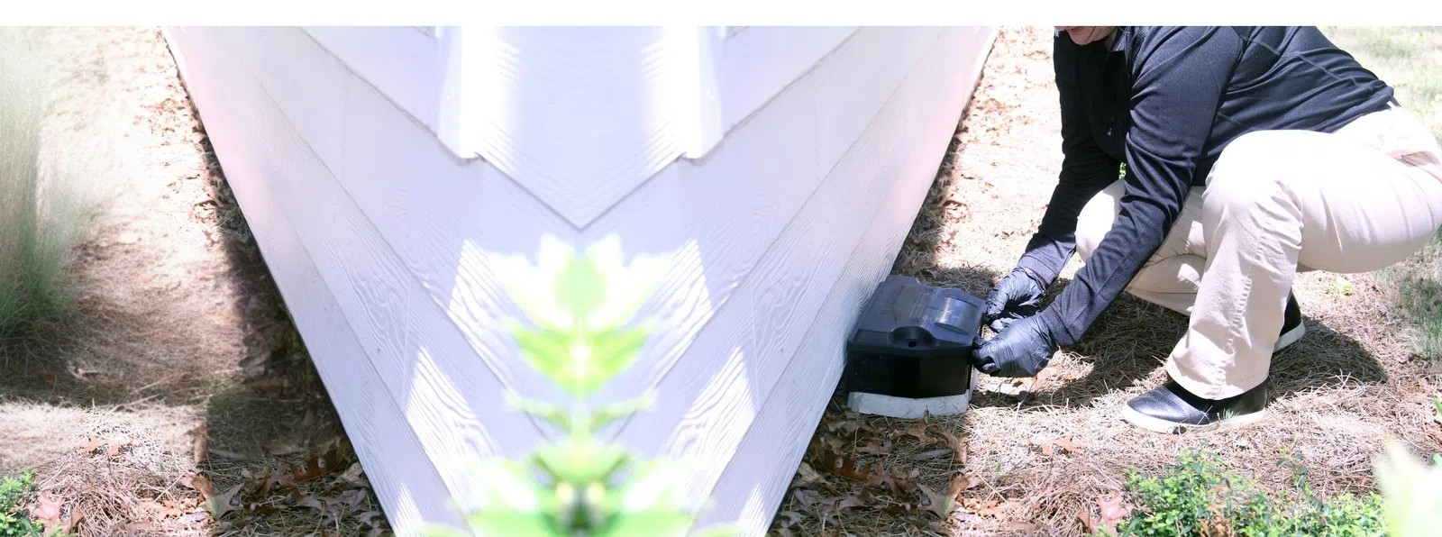 Person wearing gloves setting a black rodent bait station beside a white house exterior wall outdoors.