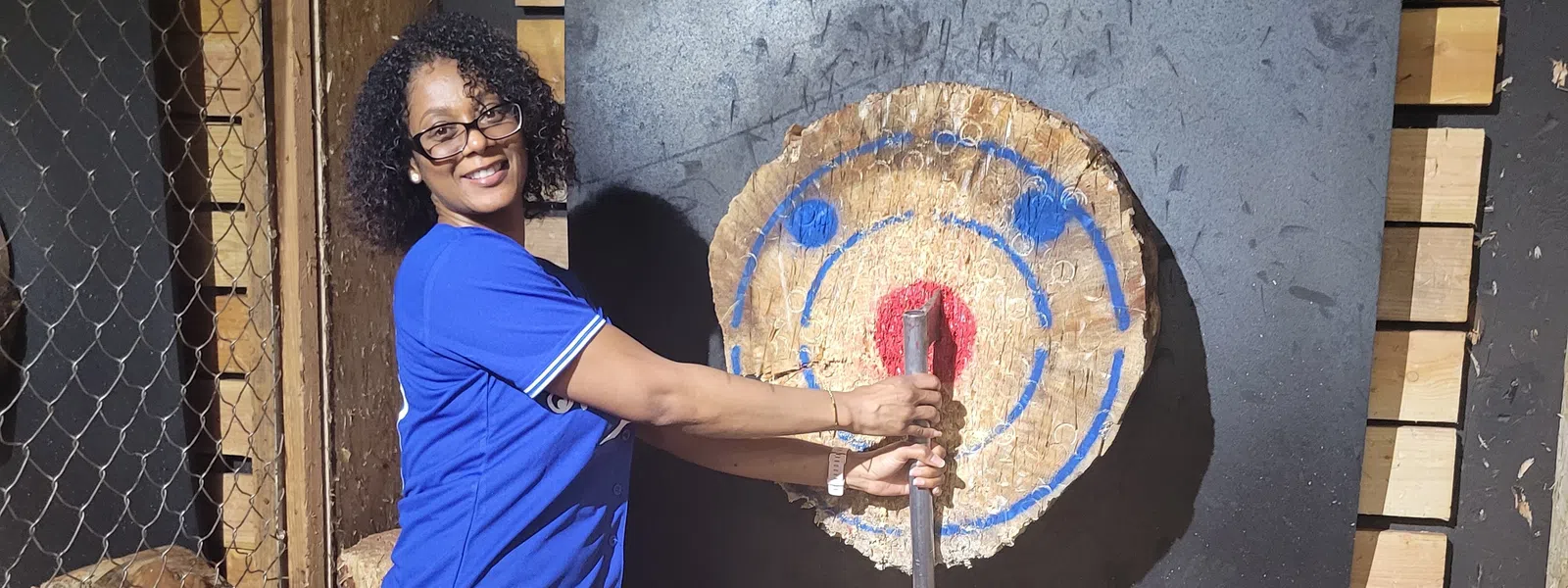 Woman in blue shirt smiling while holding an axe in front of a chopped wood target for axe throwing.