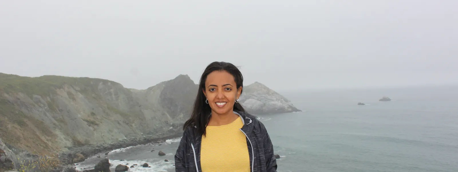 Smiling woman in yellow shirt and black jacket standing on a rocky coastline with ocean and cliffs in the background.